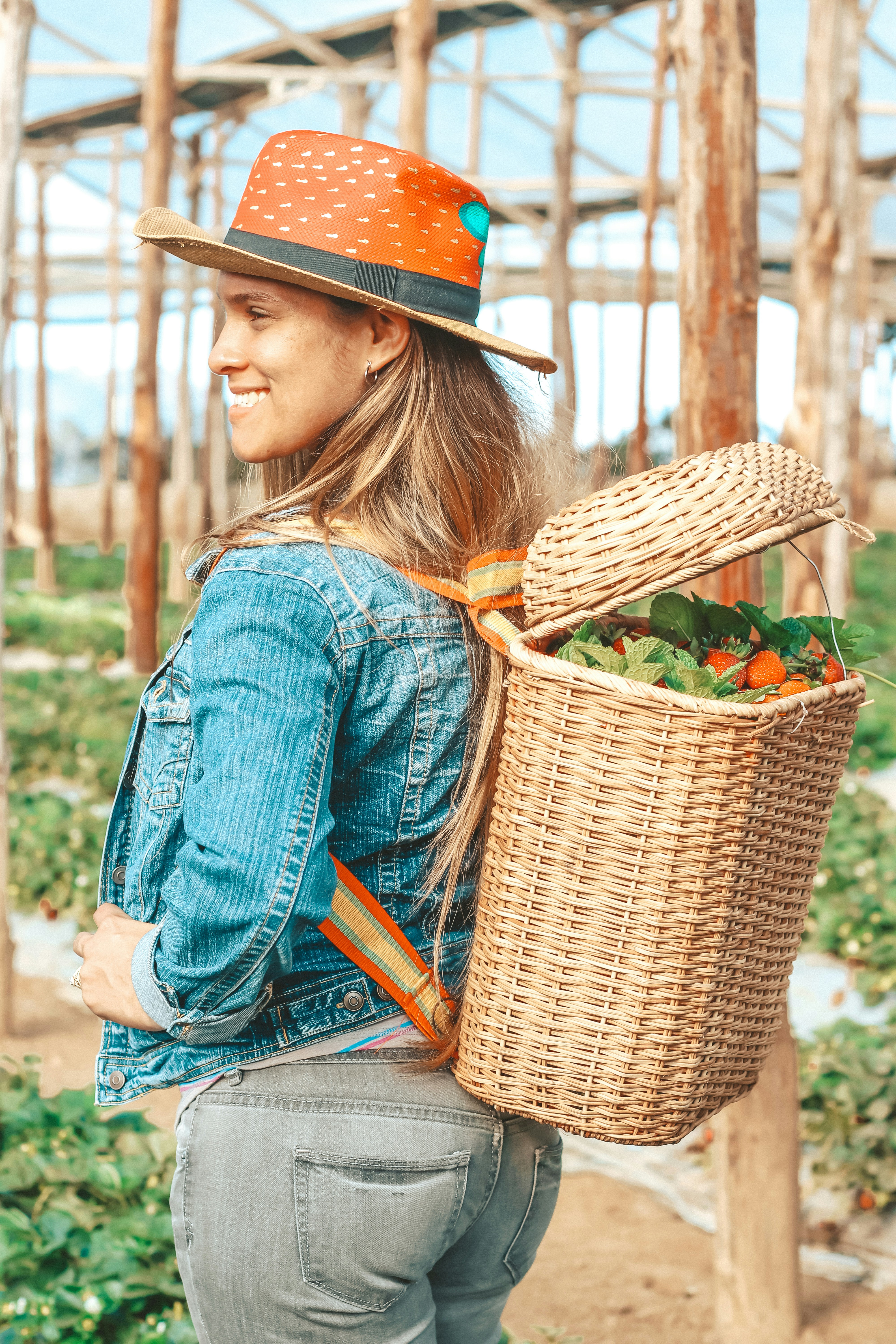 A person with long hair wearing a wide-brimmed hat, a denim jacket, and jeans stands in a greenhouse. They have a woven basket full of colorful vegetables or fruits on their back, and the setting suggests a farm or garden environment with wooden supports and greenery.