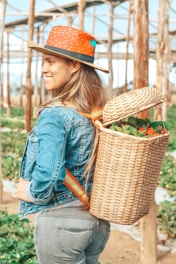 A person with long hair wearing a wide-brimmed hat, a denim jacket, and jeans stands in a greenhouse. They have a woven basket full of colorful vegetables or fruits on their back, and the setting suggests a farm or garden environment with wooden supports and greenery.