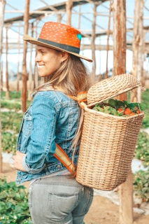 A person with long hair wearing a wide-brimmed hat, a denim jacket, and jeans stands in a greenhouse. They have a woven basket full of colorful vegetables or fruits on their back, and the setting suggests a farm or garden environment with wooden supports and greenery.