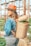 A person with long hair wearing a wide-brimmed hat, a denim jacket, and jeans stands in a greenhouse. They have a woven basket full of colorful vegetables or fruits on their back, and the setting suggests a farm or garden environment with wooden supports and greenery.