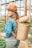 A person with long hair wearing a wide-brimmed hat, a denim jacket, and jeans stands in a greenhouse. They have a woven basket full of colorful vegetables or fruits on their back, and the setting suggests a farm or garden environment with wooden supports and greenery.