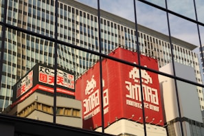 Reflections of a cityscape are visible on a large glass building facade, showcasing a prominent red building with bold white lettering and graphics. The reflection includes geometric patterns from the adjacent high-rise with vertical lines. The composition highlights architecture and urban textures.