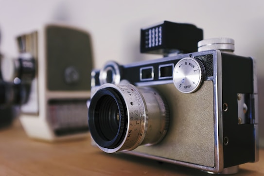 A vintage film camera with silver detailing and a lens with engraved markings on the front. The camera rests on a wooden surface, with another blurred electronic device in the background.