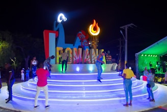 A vibrant nighttime scene featuring people gathering around a large, illuminated sign that spells 'ROMANA'. The setting includes a stage with various colors and a decorative torch-like flame. Several people are interacting, taking photos, and enjoying the atmosphere.