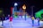 A vibrant nighttime scene featuring people gathering around a large, illuminated sign that spells 'ROMANA'. The setting includes a stage with various colors and a decorative torch-like flame. Several people are interacting, taking photos, and enjoying the atmosphere.