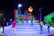 A vibrant nighttime scene featuring people gathering around a large, illuminated sign that spells 'ROMANA'. The setting includes a stage with various colors and a decorative torch-like flame. Several people are interacting, taking photos, and enjoying the atmosphere.