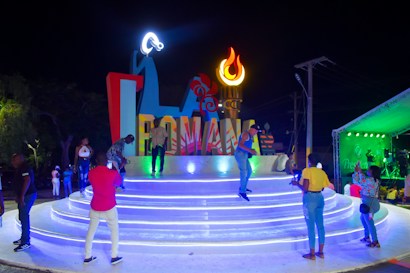 A vibrant nighttime scene featuring people gathering around a large, illuminated sign that spells 'ROMANA'. The setting includes a stage with various colors and a decorative torch-like flame. Several people are interacting, taking photos, and enjoying the atmosphere.