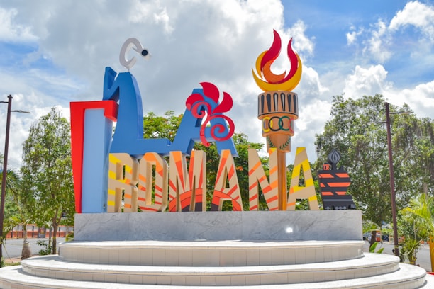 A colorful, large sign with stylized letters spelling out 'ROMANA' on a multi-tiered stone platform. Decorative elements include a flame, abstract sculptures, and an Olympic symbol. Lush green trees and a partly cloudy blue sky fill the background.