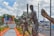A series of bronze statues depicting baseball players in action. In the background, a colorful sign reads 'ROMANA', surrounded by lush greenery and bright orange borders. The scene is set on a pedestrian pathway with additional urban elements such as vehicles and storefronts in the distance.