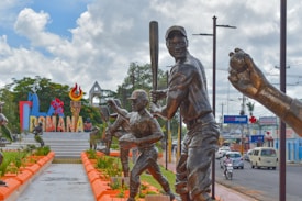 A series of bronze statues depicting baseball players in action. In the background, a colorful sign reads 'ROMANA', surrounded by lush greenery and bright orange borders. The scene is set on a pedestrian pathway with additional urban elements such as vehicles and storefronts in the distance.