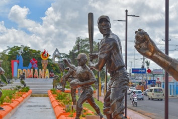 A series of bronze statues depicting baseball players in action. In the background, a colorful sign reads 'ROMANA', surrounded by lush greenery and bright orange borders. The scene is set on a pedestrian pathway with additional urban elements such as vehicles and storefronts in the distance.
