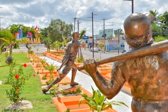 Bronze statues of baseball players swinging bats are arranged in a landscaped garden area with neatly trimmed plants and red flowers. A colorful sign in the background features large letters and symbols, along with a torch design. The scene is bordered by a street with power lines and buildings visible in the distance.