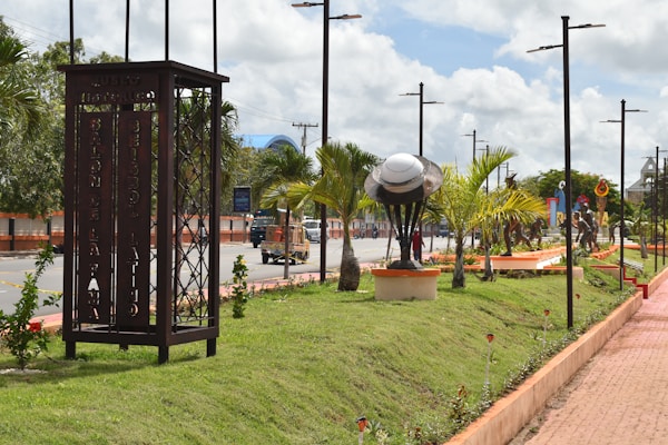 A landscaped urban park features a row of palm trees alongside a paved walkway. A metal sculpture resembling a large cap or bowl is mounted on a pedestal, surrounded by lush green grass and small plants. Tall streetlights line the walkway, and a sign with the text 'Museo Historico' is visible. Several statues of figures are placed along the path, adding an artistic touch to the scene. The sky is partly cloudy, suggesting a bright day.