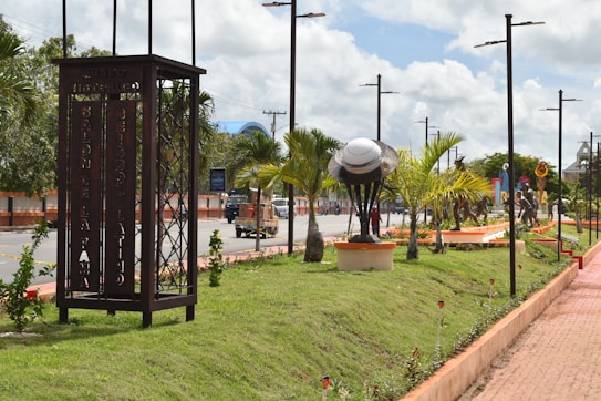 A landscaped urban park features a row of palm trees alongside a paved walkway. A metal sculpture resembling a large cap or bowl is mounted on a pedestal, surrounded by lush green grass and small plants. Tall streetlights line the walkway, and a sign with the text 'Museo Historico' is visible. Several statues of figures are placed along the path, adding an artistic touch to the scene. The sky is partly cloudy, suggesting a bright day.