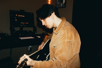 A person with dark hair is playing an acoustic guitar in a dimly lit room. The person is wearing a tan corduroy jacket and is focused on the guitar. In the background, there is a computer monitor with the text 'Liked Songs' visible, along with a keyboard and speakers.