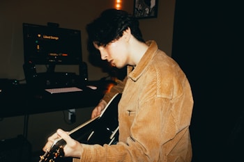 A person with dark hair is playing an acoustic guitar in a dimly lit room. The person is wearing a tan corduroy jacket and is focused on the guitar. In the background, there is a computer monitor with the text 'Liked Songs' visible, along with a keyboard and speakers.