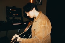 A person with dark hair is playing an acoustic guitar in a dimly lit room. The person is wearing a tan corduroy jacket and is focused on the guitar. In the background, there is a computer monitor with the text 'Liked Songs' visible, along with a keyboard and speakers.