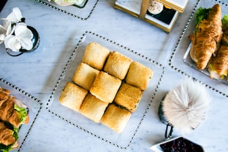 A variety of pastries and sandwiches are neatly arranged on a marble table. A square plate in the center holds baked squares with a golden crust. To the side, croissants are filled with lettuce and deli meats. There is a small, covered dish with dark fruit preserves, a decorative flower vase, and a furry decorative object.