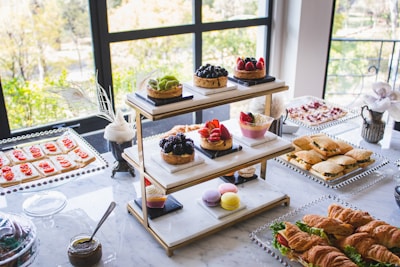 An elegantly arranged table features a variety of desserts and snacks, including assorted pastries, fruit tarts, macarons, sandwiches, croissants, and crackers with toppings. The display is set against large windows, allowing natural light to illuminate the spread, enhancing the colors and textures of the food items.