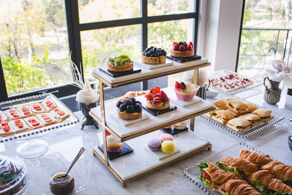 An elegantly arranged table features a variety of desserts and snacks, including assorted pastries, fruit tarts, macarons, sandwiches, croissants, and crackers with toppings. The display is set against large windows, allowing natural light to illuminate the spread, enhancing the colors and textures of the food items.