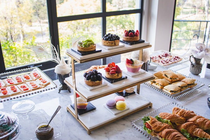 An elegantly arranged table features a variety of desserts and snacks, including assorted pastries, fruit tarts, macarons, sandwiches, croissants, and crackers with toppings. The display is set against large windows, allowing natural light to illuminate the spread, enhancing the colors and textures of the food items.