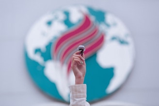 A hand is raised, holding a small gavel against a backdrop featuring an abstract representation of a globe with blue continents and a stylized red and teal flame design across it. The focus is on the hand and gavel, with the background slightly blurred.