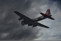 A vintage military aircraft, likely from World War II, flies through a cloudy sky. The plane's markings and insignia are visible, along with its camouflaged design and red accents on the tail and wings.