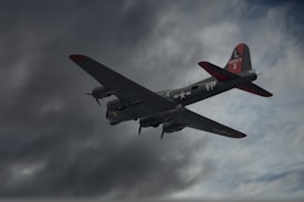 A vintage military aircraft, likely from World War II, flies through a cloudy sky. The plane's markings and insignia are visible, along with its camouflaged design and red accents on the tail and wings.