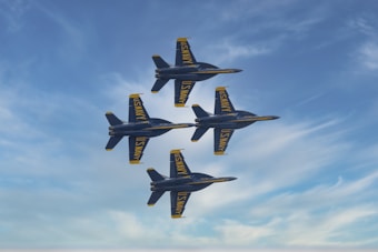 Four military fighter jets are flying in a tight diamond formation against a partly cloudy blue sky. The jets have dark blue bodies with bright yellow accents and display the words 'U.S. NAVY' on their wings and fuselage.