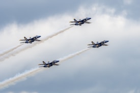 Four navy-blue fighter jets are flying in a tight formation, trailing white smoke against a backdrop of a partly cloudy sky.