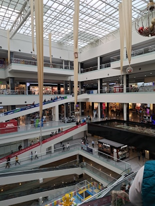 A multi-level shopping mall with various shops and stores visible across different floors. The architecture includes glass railings and large decorative fabric banners hanging from the ceiling. People are walking along the corridors and escalators, creating a bustling atmosphere. The ceiling is made of glass, allowing natural light to illuminate the interior.