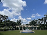 A landscaped garden features a row of tall palm trees against a vibrant blue sky with scattered white clouds. In the foreground, a circular fountain or structure is placed on a well-maintained green lawn. A building with architectural details is partially visible behind the trees.