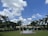 A landscaped garden features a row of tall palm trees against a vibrant blue sky with scattered white clouds. In the foreground, a circular fountain or structure is placed on a well-maintained green lawn. A building with architectural details is partially visible behind the trees.