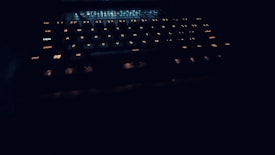 A dark setting with a keyboard that has illuminated keys, glowing softly in a warm light. The focus is on the keys, which are the main visible element in the dim lighting, creating a mysterious and high-tech vibe.