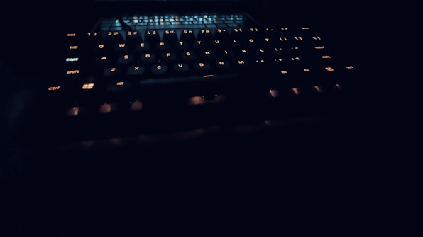A dark setting with a keyboard that has illuminated keys, glowing softly in a warm light. The focus is on the keys, which are the main visible element in the dim lighting, creating a mysterious and high-tech vibe.