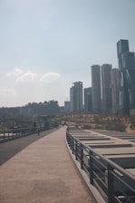 A modern urban landscape features a long, paved walkway flanked by metal railings. In the background, a cluster of contemporary skyscrapers with unique architectural designs rises into the clear sky. The left side of the image shows a park-like area with sparse greenery and trees.