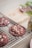 Freshly baked dark red cookies with white chocolate chips rest on a cooling rack. A wooden board and a small plate with green plants are partially visible in the background.