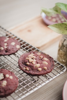 Freshly baked dark red cookies with white chocolate chips rest on a cooling rack. A wooden board and a small plate with green plants are partially visible in the background.