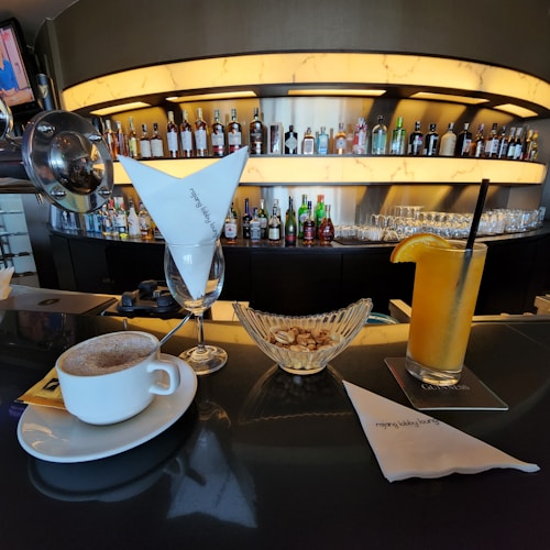 A bar setting featuring a variety of liquor bottles neatly arranged on illuminated shelves. In the foreground, there is a cup of coffee with a saucer, a glass of orange juice with a slice of orange on the rim, and a bowl of peanuts. Beside them is a glass with a white napkin, and another napkin sits on the counter with the words 'lobby lounge' printed on it.