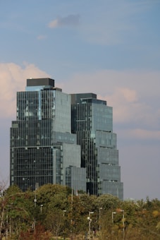 A modern, tiered skyscraper featuring reflective glass windows rises above a green landscape of trees. The sky is blue with soft, scattered clouds in the background.