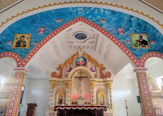 An ornate chapel interior with elaborately decorated arches and ceiling featuring religious murals with angels and saints. The central altar has detailed carvings and vibrant colors, with a cross and candleholder visible.