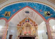 An ornate chapel interior with elaborately decorated arches and ceiling featuring religious murals with angels and saints. The central altar has detailed carvings and vibrant colors, with a cross and candleholder visible.