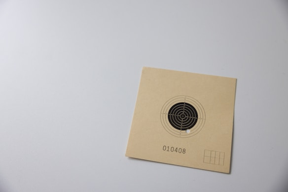 A paper card featuring a black and white concentric circle bullseye target with a single hole in it. There is a number 010408 printed below the target, and the card is laid on a light gray surface.