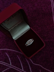 A jewelry box with a red exterior and velvet interior contains a diamond-studded ring. The ring has a distinctive design, and the box is positioned on a purple, textured fabric background.