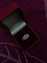 A jewelry box with a red exterior and velvet interior contains a diamond-studded ring. The ring has a distinctive design, and the box is positioned on a purple, textured fabric background.