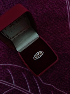A jewelry box with a red exterior and velvet interior contains a diamond-studded ring. The ring has a distinctive design, and the box is positioned on a purple, textured fabric background.