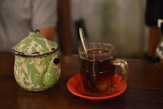 A rustic earthenware mug with a vintage-inspired design sitting on a wooden table beside a steaming teapot.