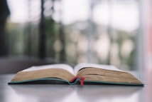 An open book lies flat on a reflective surface with a red bookmark hanging down from the center. The background is blurred, suggesting an out-of-focus outdoor view.