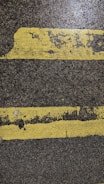 Two parallel yellow lines, worn and cracked, are painted on a rough, dark asphalt surface. The texture of the pavement and paint is visible, showing signs of wear and tear.