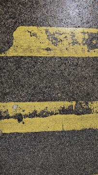 Two parallel yellow lines, worn and cracked, are painted on a rough, dark asphalt surface. The texture of the pavement and paint is visible, showing signs of wear and tear.
